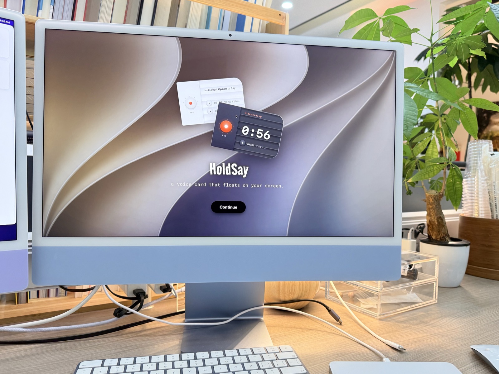 A blue iMac on an office desk with bookshelves and a plant behind it, its screen showing the HoldSay card and website.
